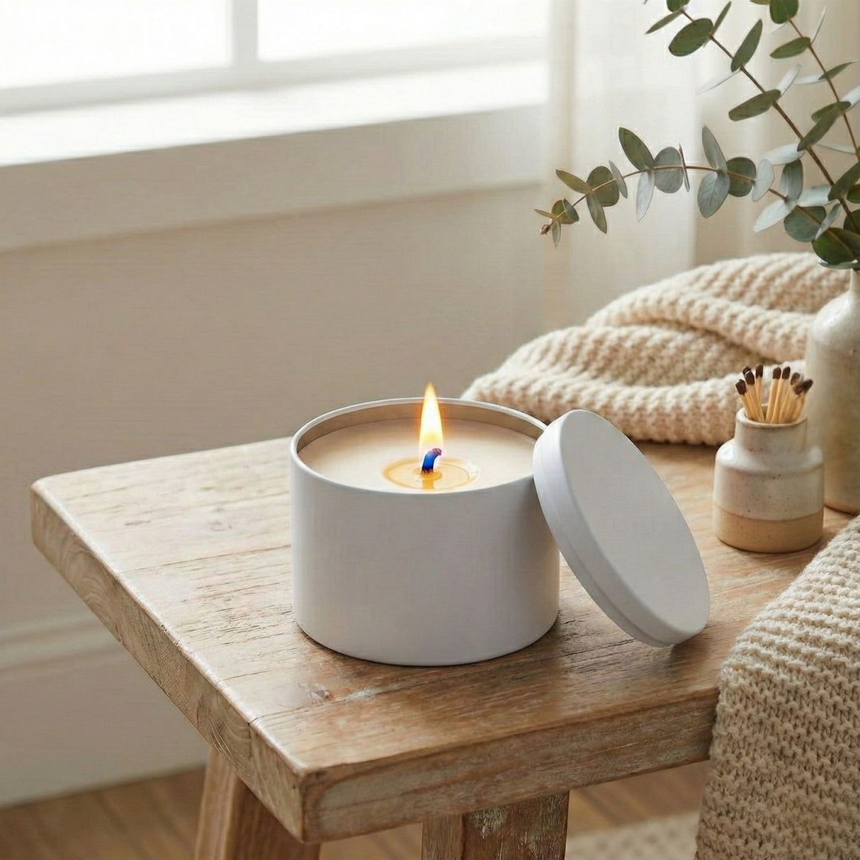 White candle in a tin with a lit wick on a wooden table, surrounded by decorative items.