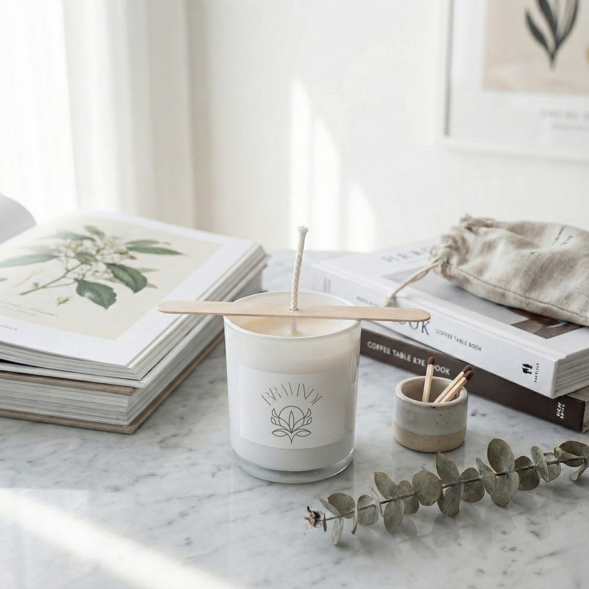 White candle with wooden wick holder on a marble surface with books and eucalyptus branch.