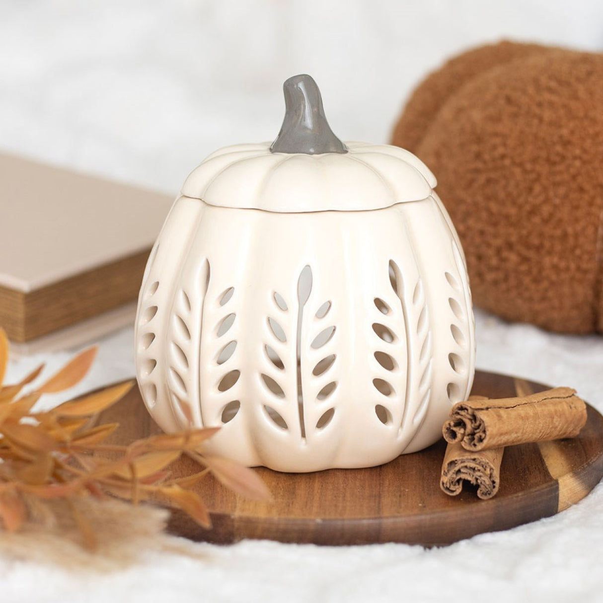 White ceramic pumpkin-shaped lantern with cut-out designs on a wooden board with cinnamon sticks and leaves.