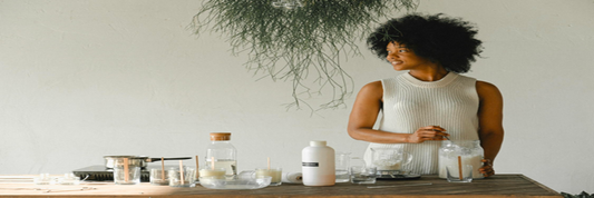 A smiling woman stirring a jar of wax with candle-making supplies laid out. Sourced from Pexels