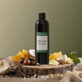 Bottle of 'Chestnuts & Coconuts' on a wooden stand with vanilla beans and oranges on a green background