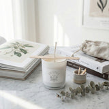 White candle with wooden wick holder on a marble surface with books and eucalyptus branch.