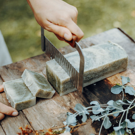 Hand using a knife to cut soap bars on a wooden surface with greenery in the background
