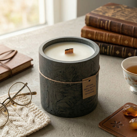 Candle in a dark container on a table with books, glasses, and a tea cup in the background.